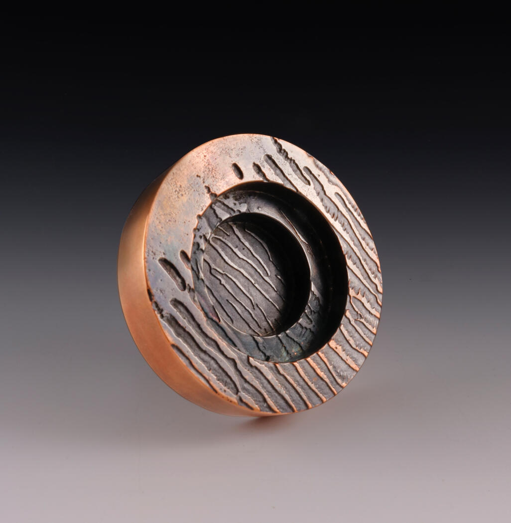 A round copper brooch with a dark, textured center and engraved, flowing lines resembling erosion or topography. The design evokes a planetary surface, blending organic patterns with metallic tones in a sculptural, abstract form.