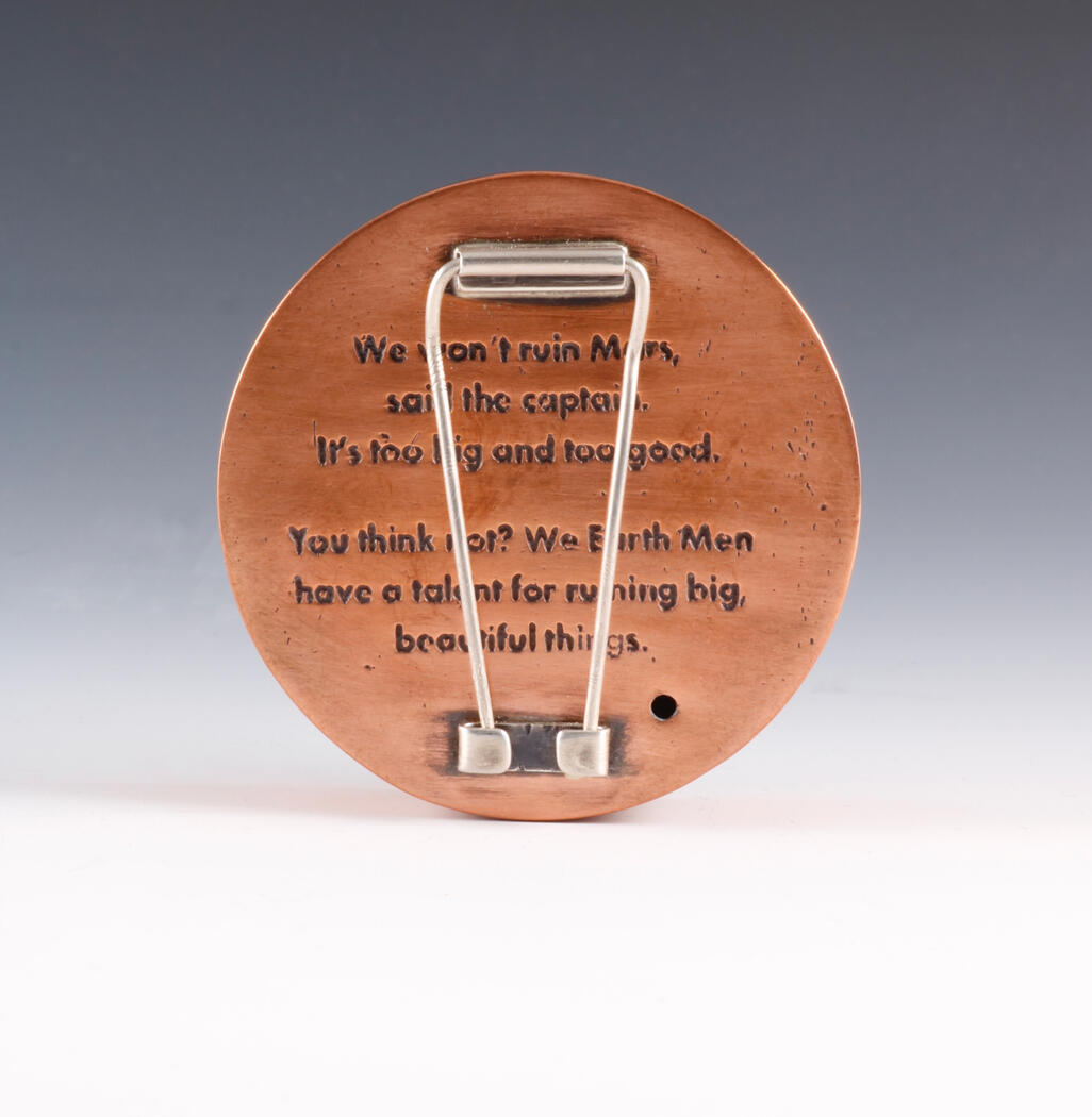 The back of the round, copper brooch is engraved with text and fitted with a silver pin clasp. The inscription references Mars and human destruction, blending jewelry craftsmanship with a reflective message about humanity’s impact on beauty.