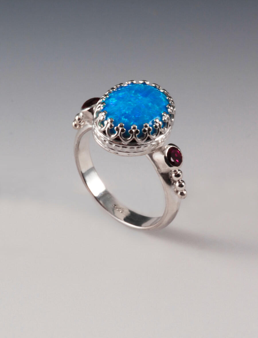 Charlie Ring 1 by Charles Mirkin A silver ring featuring a large, vibrant blue gemstone set in an ornate crown-like bezel. Two small deep red stones accent the sides, with delicate silver bead detailing on a smooth, polished band against a neutral background.